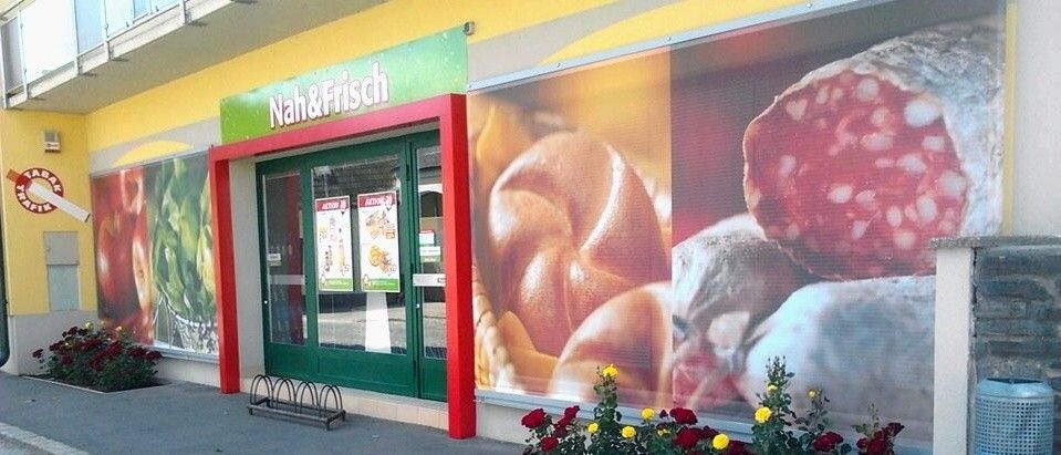 Außenansicht eines Lebensmittelgeschäfts mit roten Blumen und einer grünen Tür. Der Name des Geschäfts lautet Nah&Frisch.