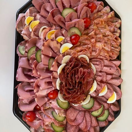 Ein schwarzer Teller mit einer Auswahl an geschnittenen Fleischsorten, einschließlich Schinken, Salami und Prosciutto, in einer Blumenform mit Gurkenscheiben und Kirschtomaten angeordnet.