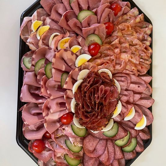 Ein schwarzer Teller mit einer Auswahl an geschnittenen Fleischsorten, einschließlich Schinken, Salami und Prosciutto, in einer Blumenform mit Gurkenscheiben und Kirschtomaten angeordnet.