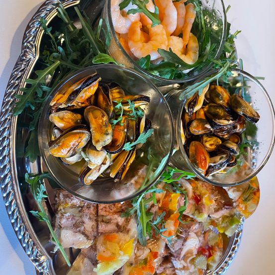 Eine silberne Platte mit drei Glasbowlen gefüllt mit Muscheln und Garnelen, garniert mit grünen Blättern, und einem farbenfrohen Gericht aus gemischtem Meeresfrüchten.