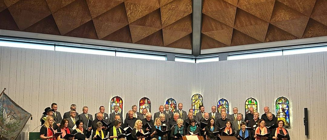 Ein Chor singt in einer Kirche mit einer Holzdecke und grünen Schals. Das Publikum sitzt vor ihnen in Stühlen.