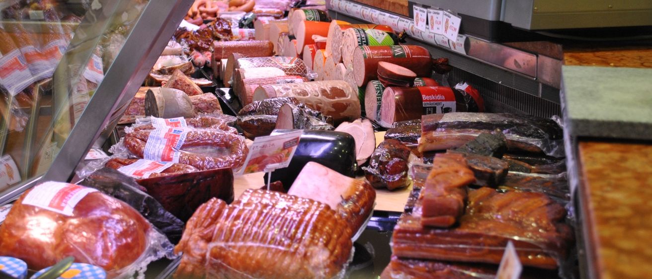 Ein Delikatessen-Schalter mit verschiedenen Würsten und Fleischsorten, einschließlich Frankfurter Würsten, Salami und gereiftem Fleisch, ordentlich angeordnet und bepreist. Der Schalter ist mit einer Glasvitrine abgedeckt, und im Hintergrund befinden sich eine Waage und eine Kasse.