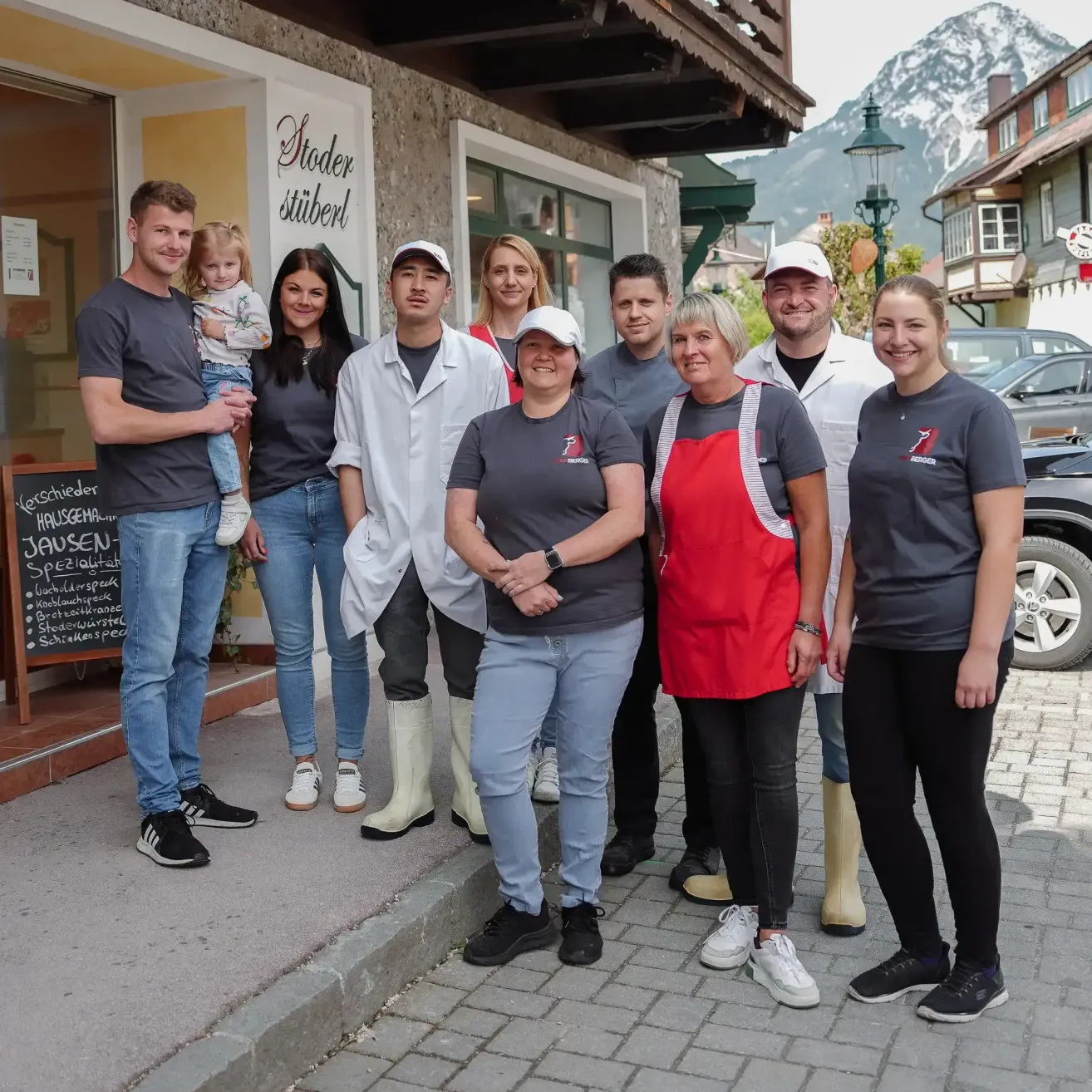 Eine Gruppe von Menschen in passenden grauen und roten T-Shirts posiert vor einem Gebäude mit dem Schild 'Stoeder'. Sie stehen auf einem gepflasterten Bürgersteig mit Bergen im Hintergrund.