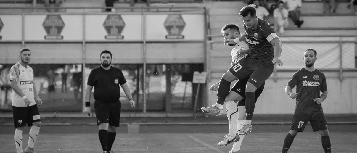 Ein Fußballspiel ist im Gange. Zwei Spieler sind in der Luft, einer versucht den Ball zu köpfen. Zuschauer schauen von den Tribünen zu, und ein Schiedsrichter steht links.