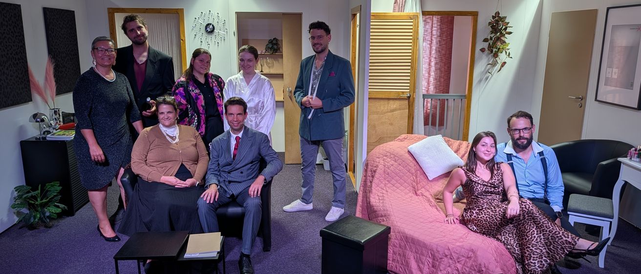 Eine Gruppe von Menschen in verschiedenen Outfits posiert für ein Foto auf einer Bühne. Einige sitzen auf einem Sofa, andere stehen. Es gibt zwei Tische und eine Aufbewahrungsbox auf der Bühne.