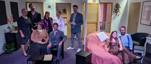 Eine Gruppe von Menschen in verschiedenen Outfits posiert für ein Foto auf einer Bühne. Einige sitzen auf einem Sofa, andere stehen. Es gibt zwei Tische und eine Aufbewahrungsbox auf der Bühne.