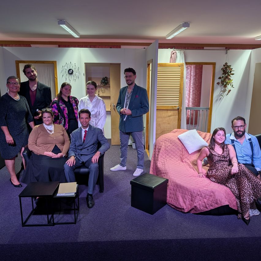 Eine Gruppe von Menschen in verschiedenen Kostümen posiert für ein Foto auf einer Bühnendekoration mit einem Bett, Stühlen und einem Hintergrund.