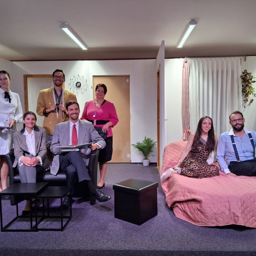 Eine Gruppe von Menschen in formeller Kleidung posiert für ein Foto in einem Raum mit Bühnenaufbau, mit einer Couch, Stühlen und einer kleinen Pflanze.