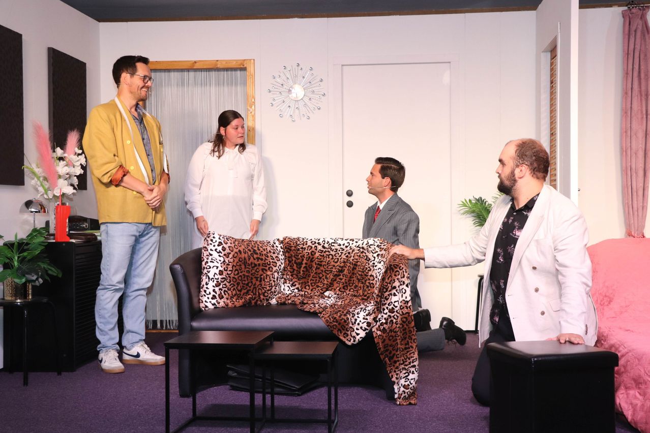 Fünf Personen sind in einem Raum, eine Person hält eine Leopardendecke. Eine Couch mit Leopardenkissen steht in der Mitte. Ein Mann im Anzug kniet, und ein Mann im weißen Mantel hält die Decke.