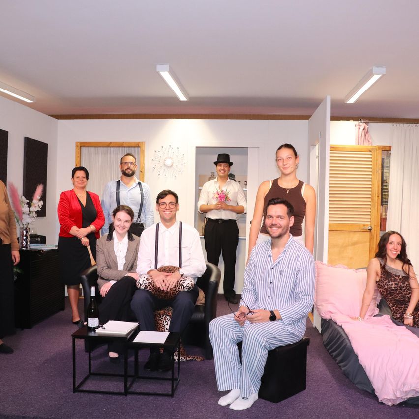 A group of people are posing for a photo in a room with white walls and purple carpet. Some are sitting on a couch, others standing. One person holds a bouquet of flowers.