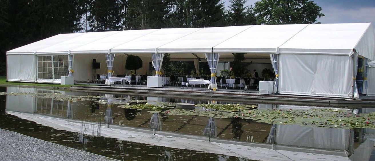Ein großes weißes Zelt für eine Veranstaltung, reflektiert in einem Teich mit Seerosen. Bäume umgeben das Gebiet, und Tische und Stühle sind im Inneren angeordnet.