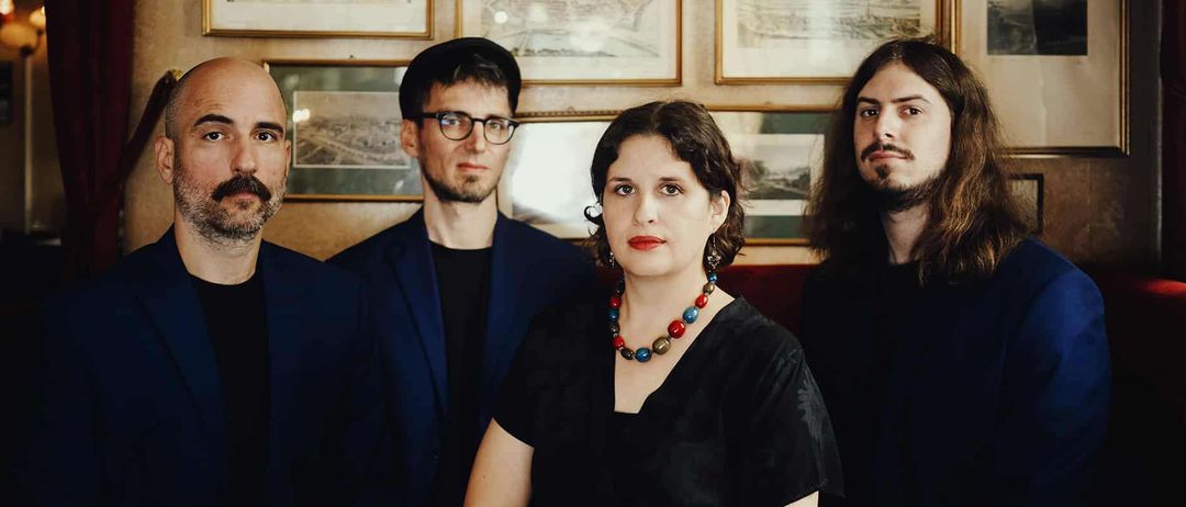 Vier Menschen in formeller Kleidung sitzen gemeinsam auf einem Sofa und blicken in die Kamera. Sie sind von mehreren gerahmten Bildern an einer Wand umgeben.