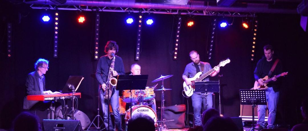 Eine Band tritt auf der Bühne unter bunten Lichtern auf. Ein Saxophonist steht in der Mitte, während ein Gitarrist und ein Schlagzeuger hinter ihm spielen. Das Publikum sitzt davor und genießt die Musik.
