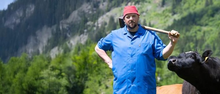 Ein Mann steht in einem Grasfeld mit zwei Kühen, hält einen Vorschlaghammer und trägt einen blauen Mantel und einen roten Hut. Hinter ihm befindet sich ein bewaldeter Hügel.