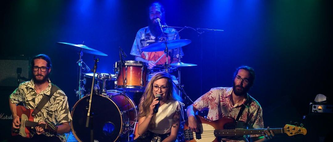 Eine Band tritt auf der Bühne auf, mit einem Sänger, der ein Mikrofon hält, einem Gitarristen und einem Schlagzeuger. Die Bühne ist mit blauer Beleuchtung beleuchtet.