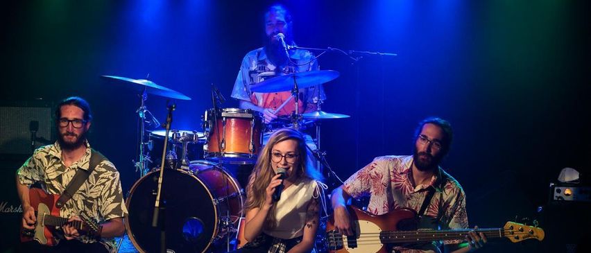 Eine Band tritt auf der Bühne auf, mit einem Sänger, der ein Mikrofon hält, einem Gitarristen und einem Schlagzeuger. Die Bühne ist mit blauer Beleuchtung beleuchtet.