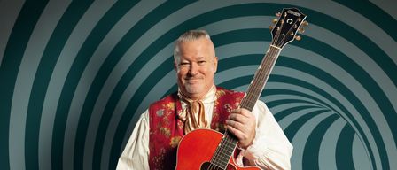 Ein Mann in einem Wams steht mit einer roten Gitarre und lächelt in die Kamera. Die Gitarre hat einen schwarzen Plektrumhalter und der Hintergrund ist ein grünes Spiralmuster.