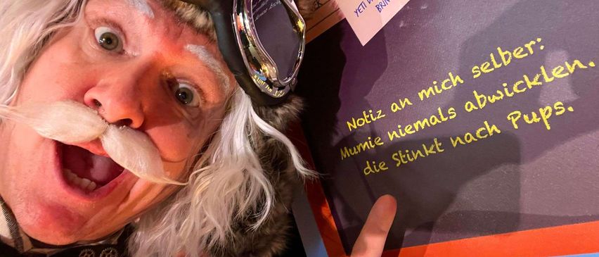 Ein Mann mit Schnurrbart, Schutzbrille und Pelzmütze zeigt mit dem Finger auf ein Schild mit gelbem Text auf dunklem Hintergrund. Das Schild lautet: 'Beachte, dass ich nie nach Mumie rieche.'