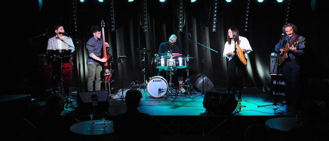 Eine Band tritt auf der Bühne auf, mit Scheinwerfern und schwarzem Hintergrund. Die Band besteht aus einem Schlagzeuger, einem Bassisten, einem Gitarristen und einem Sänger. Das Publikum sitzt vor der Bühne.