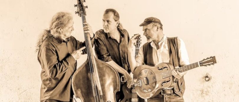 Drei Männer mit Instrumenten stehen vor einer weißen Wand. Der Mann auf der linken Seite hält eine große Bassgitarre. Der Mann in der Mitte hält eine Gitarre und einen Resonator. Der Mann auf der rechten Seite trägt einen Hut.