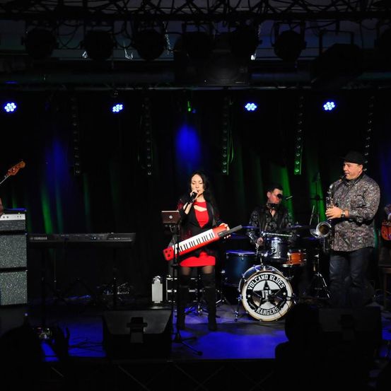 Eine Band tritt auf einer Bühne mit Scheinwerfern auf. Eine Frau in einem roten Kleid singt und hält eine rote Tastatur. Ein Mann in einem Hut spielt Saxophon. Dahinter steht ein Schlagzeug mit einem Logo.