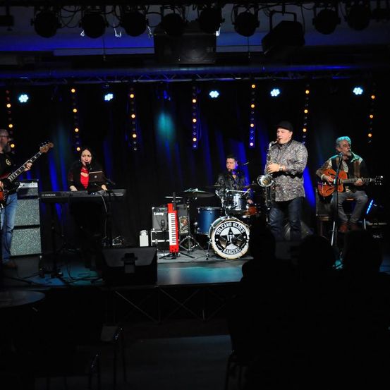 Eine Band spielt auf der Bühne mit einem Schlagzeug, einem Saxophonisten und einem Keyboarder. Das Publikum ist im Hintergrund dunkel.