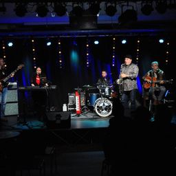 Eine Band spielt auf der Bühne mit einem Schlagzeug, einem Saxophonisten und einem Keyboarder. Das Publikum ist im Hintergrund dunkel.