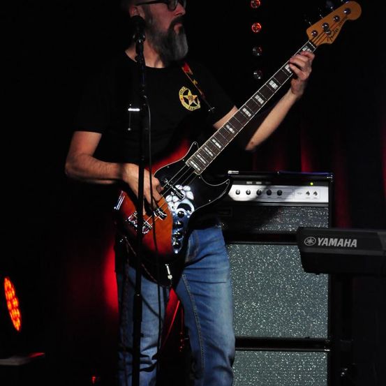 Ein bärtiger Mann spielt eine E-Gitarre auf der Bühne, beleuchtet von roten Lichtern, mit einem Yamaha-Keyboard dahinter.