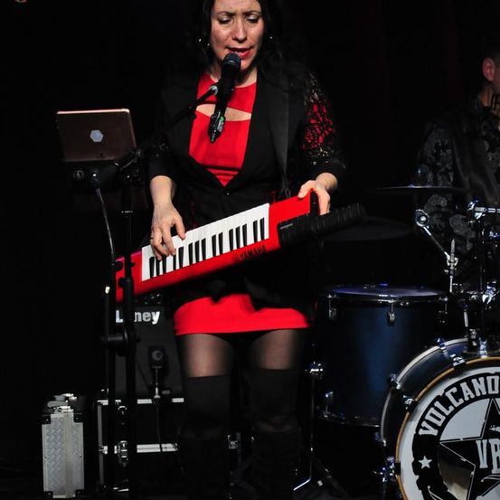 Eine Frau tritt auf der Bühne auf, hält ein rotes Keyboard und singt in ein Mikrofon. Ein Schlagzeug mit einem Sternenlogo ist hinter ihr zu sehen.