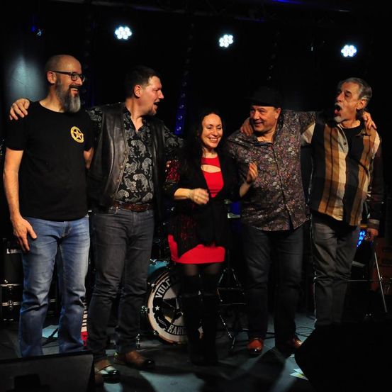 Eine Band posiert für ein Foto auf der Bühne mit Scheinwerfern im Hintergrund, ein Mitglied hält ein Schlagzeug.