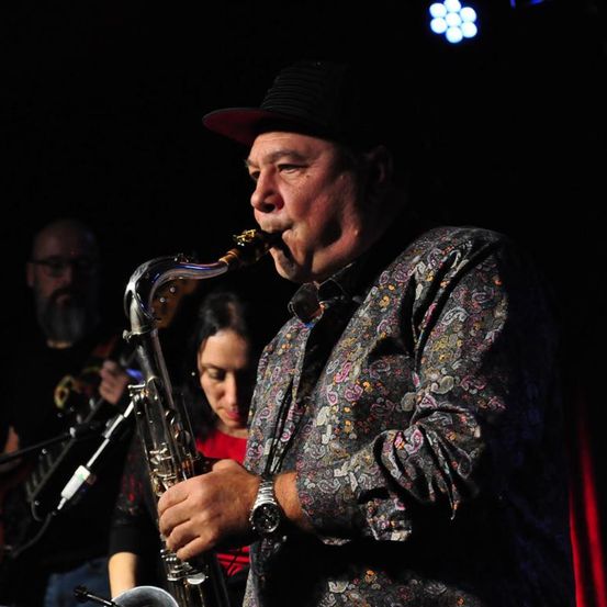 Ein älterer Mann spielt Saxophon in einem schwach beleuchteten Raum. Er trägt einen Hut und eine Uhr. Hinter ihm spielt eine Frau Gitarre.