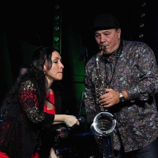 Ein Mann mit schwarzem Hut spielt Saxophon, während eine Frau in einem roten Oberteil ihn auf der Bühne ansieht.