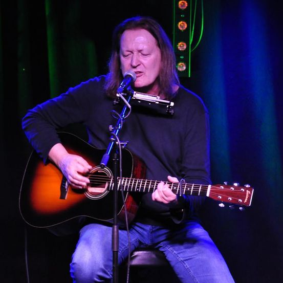Eine Person sitzt auf einem Stuhl und spielt Gitarre, während sie in ein Mikrofon singt. Er trägt ein schwarzes Langarmshirt und blaue Jeans.