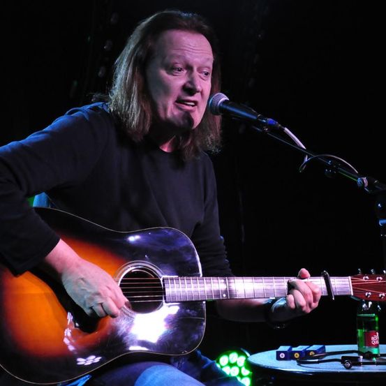 Ein Mann spielt auf der Bühne mit einer Gitarre. Er greift und singt in ein Mikrofon. Er trägt ein schwarzes Hemd. Auf dem Tisch vor ihm steht eine Flasche.