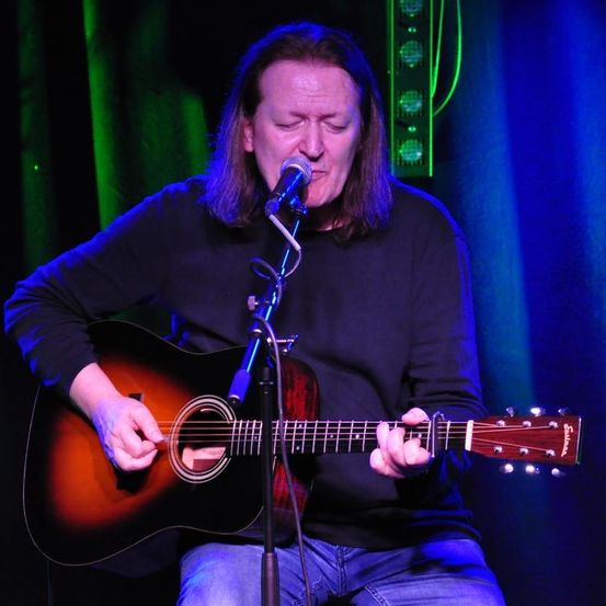 Ein Mann sitzt und spielt eine Gitarre, während er in ein Mikrofon singt. Er trägt ein schwarzes Langarmshirt und Jeans. Die Umgebung scheint eine Bühne mit blauer und grüner Beleuchtung zu sein.