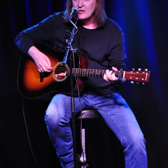 Ein Mann sitzt auf einem Hocker und spielt Gitarre. Er hält die Gitarre mit der linken Hand und zupft sie mit der rechten Hand. Er singt in ein Mikrofon, das auf einem Ständer steht. Er trägt ein schwarzes Langarmshirt und blaue Jeans.