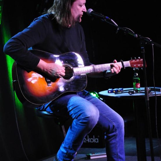 Ein Mann mit langen Haaren sitzt auf einem Hocker, spielt Gitarre und singt in ein Mikrofon. Vor ihm stehen eine grüne Flasche und ein kleiner Tisch mit einem Lautsprecher und anderen Gegenständen.