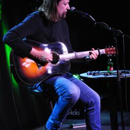 Ein Mann mit langen Haaren sitzt auf einem Hocker, spielt Gitarre und singt in ein Mikrofon. Vor ihm stehen eine grüne Flasche und ein kleiner Tisch mit einem Lautsprecher und anderen Gegenständen.