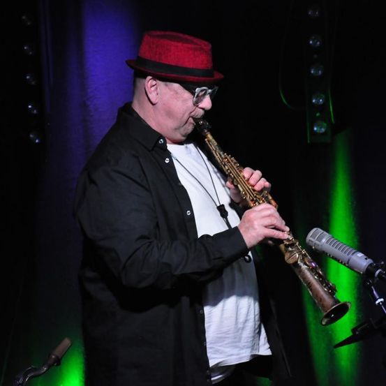 Ein Mann mit rotem Hut und Brille spielt Saxophon auf der Bühne unter grünen und lila Lichtern.
