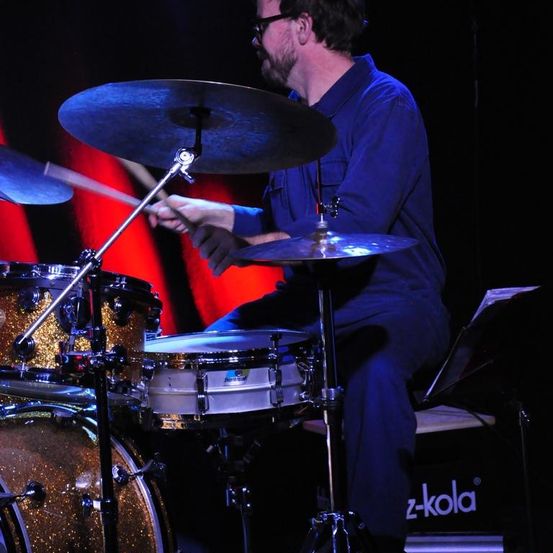 Ein Mann spielt Schlagzeug auf einer Bühne. Er trägt eine Brille und ein blaues Hemd. Das Schlagzeug hat eine Becken und einen Bassdrum.