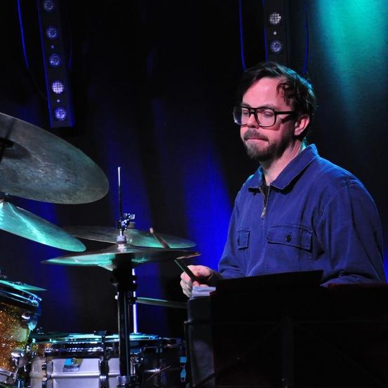 Ein Mann spielt die Drums auf der Bühne, trägt eine Brille und ein blaues Hemd. Das Drumset ist sichtbar, zusammen mit mehreren Becken und einer Spotlight hinter ihm.