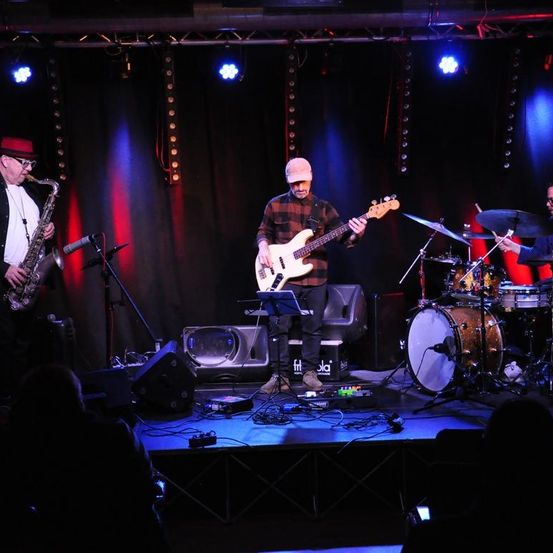 Eine Band tritt auf einer Bühne mit Scheinwerfern auf. Ein Mann spielt Saxophon, ein anderer Gitarre und ein Schlagzeuger spielt Schlagzeug.