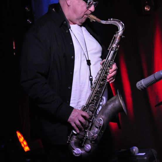 Ein älterer Mann in einem schwarzen Jackett und weißem T-Shirt spielt Saxophon vor einem roten Vorhang und mit Scheinwerfern im Hintergrund.