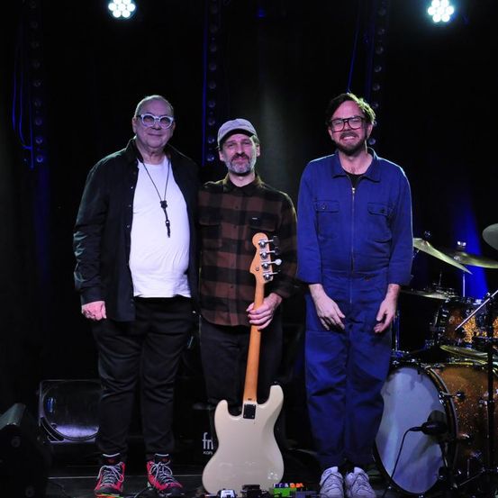 Drei Männer auf der Bühne, einer mit einer Gitarre, ein anderer mit Schlagzeug, vor einem schwarzen Vorhang und Scheinwerfern.