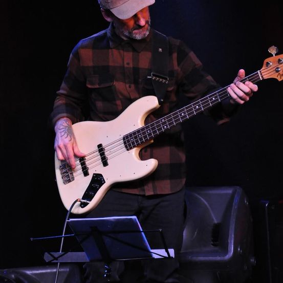 Ein Mann in einem karierten Hemd und einer Mütze spielt eine weiße Bassgitarre auf der Bühne. Er wird von einem Scheinwerfer beleuchtet.