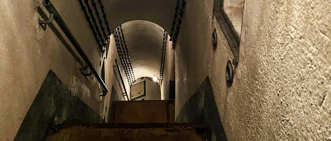 Eine schwach beleuchtete Wendeltreppe mit Metallhandläufen führt zu einem gewölbten Durchgang mit einer Tür und einem kleinen Fenster.