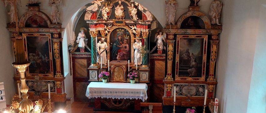 Ein Kirchenaltar mit einem zentralen Gemälde der Jungfrau Maria und des Kindes. Flankiert von Statuen, umgeben von Kerzen. Zwei Vasen mit rosa Blumen stehen davor.
