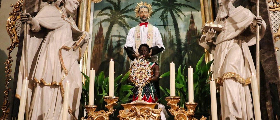 A religious altar with a golden candlestick and several candles. A priest statue stands behind a smaller statue of a person in a costume.