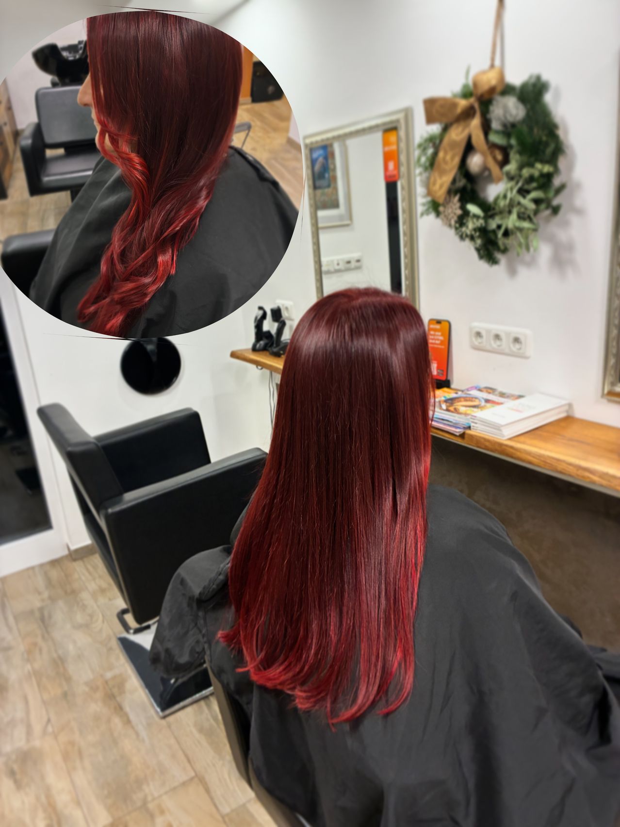 Eine Frau mit roten Haaren sitzt in einem Salon, mit einem Spiegel, der ihr Haar hinter ihr reflektiert. Der Salon hat Holzböden und eine Wand mit einem Kranz.
