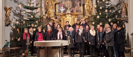 Ein Chor steht vor einem Altar in einer Kirche, geschmückt mit Weihnachtsbäumen und einem großen Gemälde dahinter.
